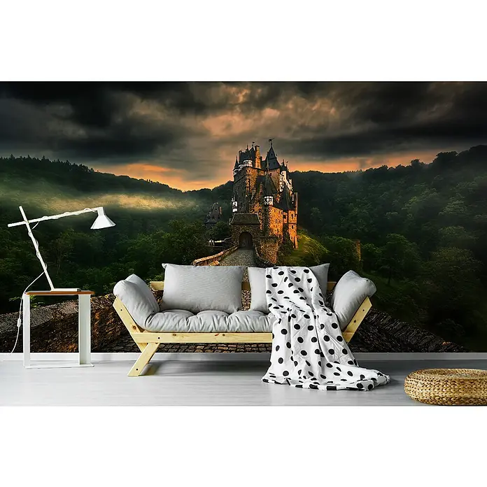 Papermoon Premium collection Fototapete Eltz I - B x H: 250 x 186 cm, Vlies - Wandbild mit einer mittelalterlichen Burg im Hintergrund, davor ein stilvolles Sofa mit Kissen und eine Lampe für eine gemütliche Atmosphäre.