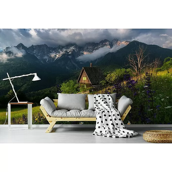 Papermoon Premium collection Fototapete Auf den Wiesen - B x H: 400 x 260 cm, Vlies - Holzsofa mit hellgrauen Kissen und weißer Polka-Dot-Decke, weißer Beistelltisch mit Lampe und Stroh-Pouf in einem Raum mit Bergwandbild.