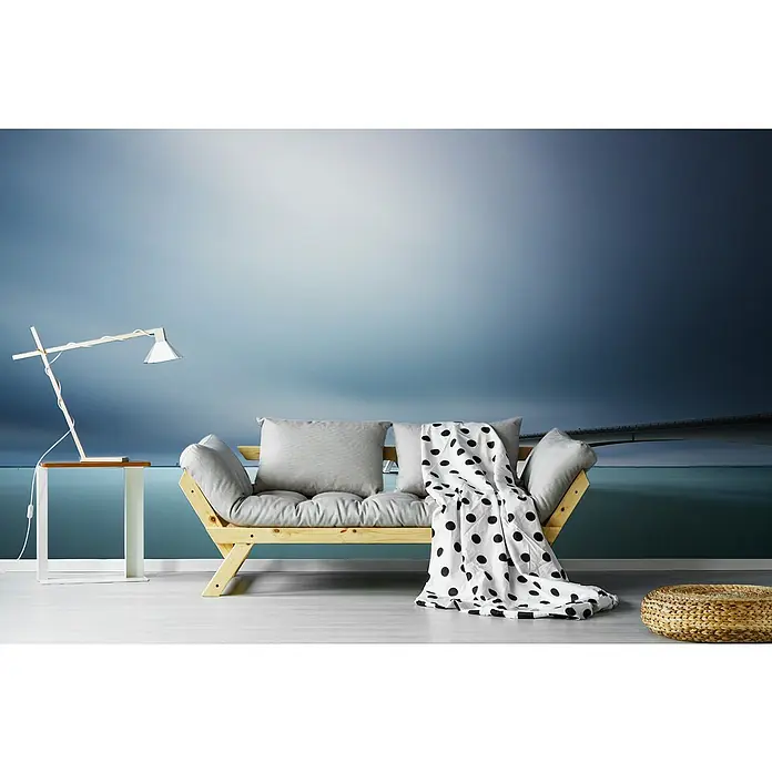 Papermoon Premium collection Fototapete Unendliche Brücke - B x H: 450 x 280 cm, Vlies - Wohnzimmer mit hellem Holzsofa, Polsterkissen und gepunkteter Decke vor einer Wand mit Brückenmotiv, dazu eine moderne Lampe und ein Beistelltisch.