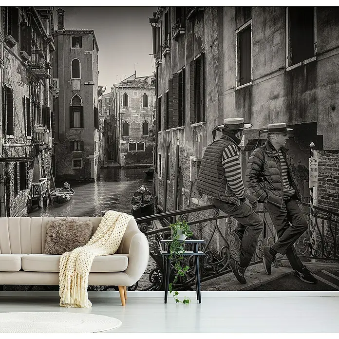 Papermoon Premium collection Fototapete Gondolieri - B x H: 200 x 149 cm, Vlies - Wandbild mit einem venezianischen Kanal, zwei Männern auf einer Brücke, einem beigen Sofa mit Kissen und einer Decke, und einem kleinen Beistelltisch.