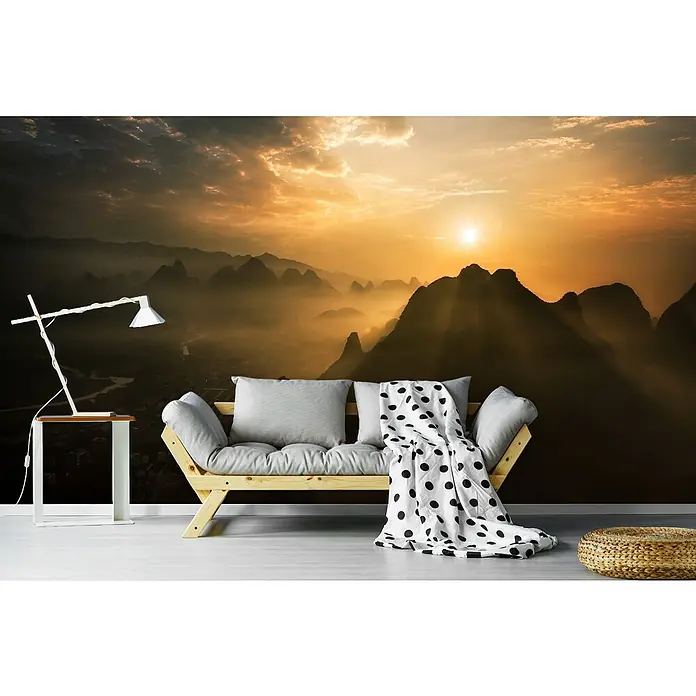 Papermoon Premium collection Fototapete Nebliger Sonnenaufgang - B x H: 450 x 280 cm, Vlies - Wohnzimmer mit Sofa, Beistelltisch und Lampe vor einer Fototapete mit Berglandschaft bei Sonnenaufgang.