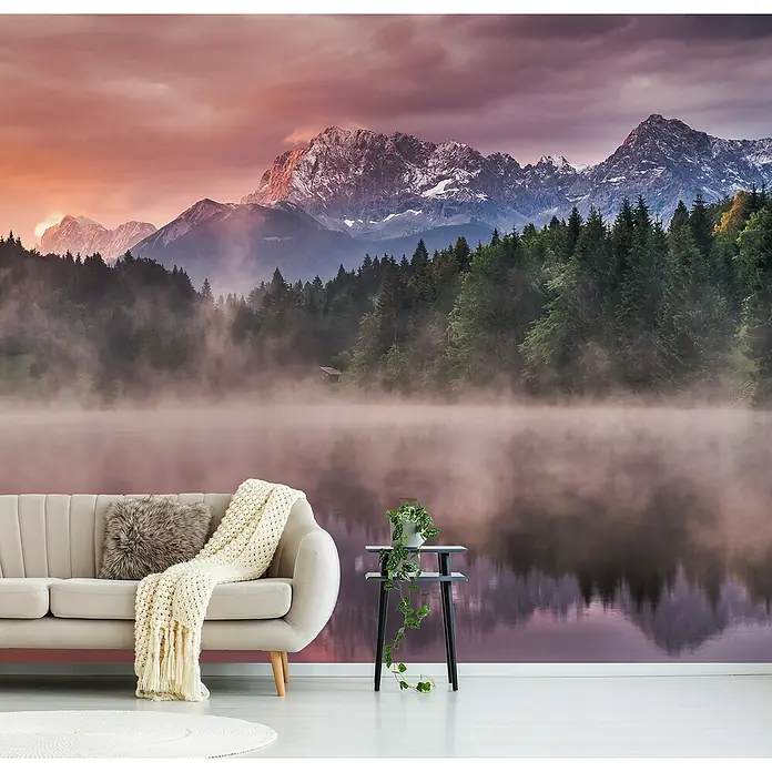 Papermoon Premium collection Fototapete Sonnenaufgang am See - B x H: 300 x 223 cm, Vlies - Ein Wohnzimmer mit einer Fototapete, die einen See, Berge und Bäume zeigt, dekoriert mit einem Sofa und einem Beistelltisch mit einer Pflanze.