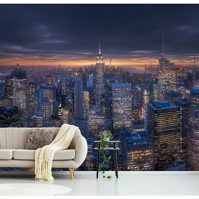 Papermoon Premium collection Fototapete Skyscraperview II - B x H: 450 x 280 cm, Vlies - Ein beiges Sofa mit flauschigem Kissen und Strickdecke, daneben eine Pflanze auf einem Beistelltisch, in einem Raum mit Stadtpanorama-Wandbild.