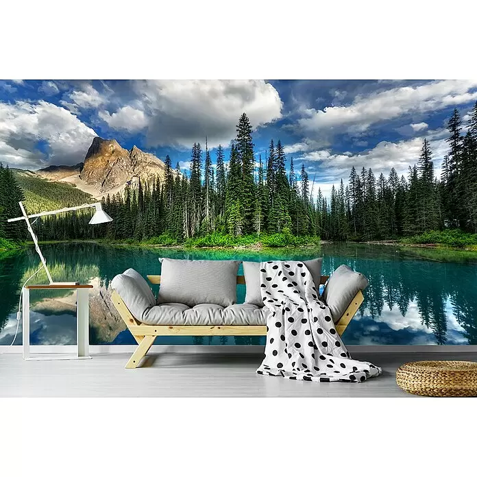 Papermoon Premium collection Fototapete Smaragd - B x H: 500 x 280 cm, Vlies - Ein Holzsofa mit grauen Kissen und gepunkteter Decke, ein weißer Beistelltisch mit Lampe und ein geflochtener Pouf in einem Raum mit Natur-Wandbild.