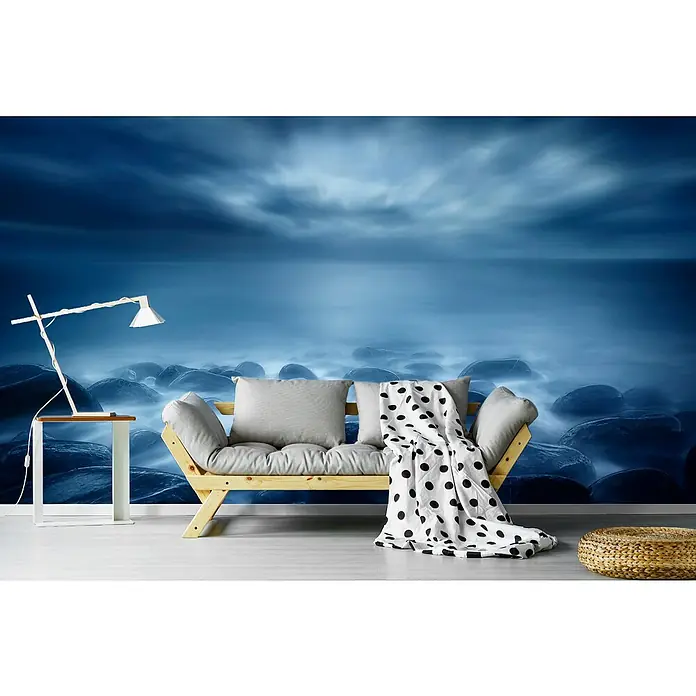 Papermoon Premium collection Fototapete Blau - B x H: 250 x 186 cm, Vlies - Wohnzimmer mit Design Lampe, hellem Holzsofa mit Kissen und einer schwarz-weißen Decke vor einer Wand mit Felsen Meerblick Motiv.