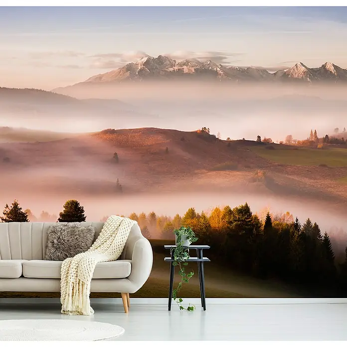 Papermoon Premium collection Fototapete Nur ein Schweigen - B x H: 450 x 280 cm, Vlies - Ein Interieur mit einer Wandtapete, die eine malerische Berglandschaft im Morgennebel zeigt, mit einem Sofa und Beistelltisch im Vordergrund.