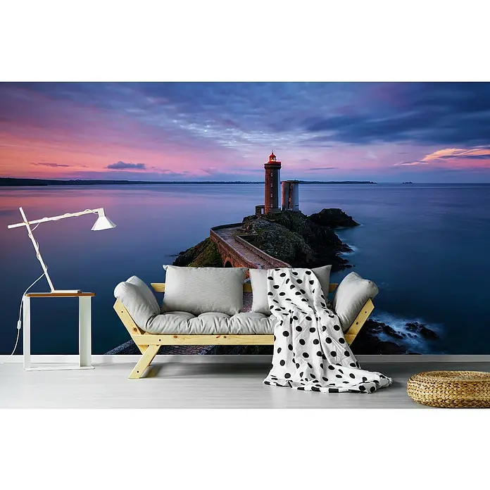 Papermoon Premium collection Fototapete Petit Minou - B x H: 450 x 280 cm, Vlies - Ein entspannender Raum mit einer Fototapete, die einen Leuchtturm bei Sonnenuntergang zeigt, mit Sofa, Beistelltisch und einem runden Hocker im Zimmer.