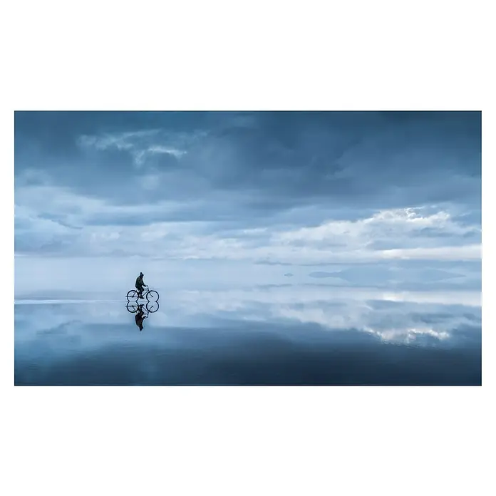 Papermoon Premium collection Fototapete In den Himmel gehen - B x H: 350 x 260 cm, Vlies - Ein einsamer Radfahrer, dessen Spiegelbild sich auf der ruhigen Wasseroberfläche widerspiegelt, unter einem wolkenverhangenen Himmel.