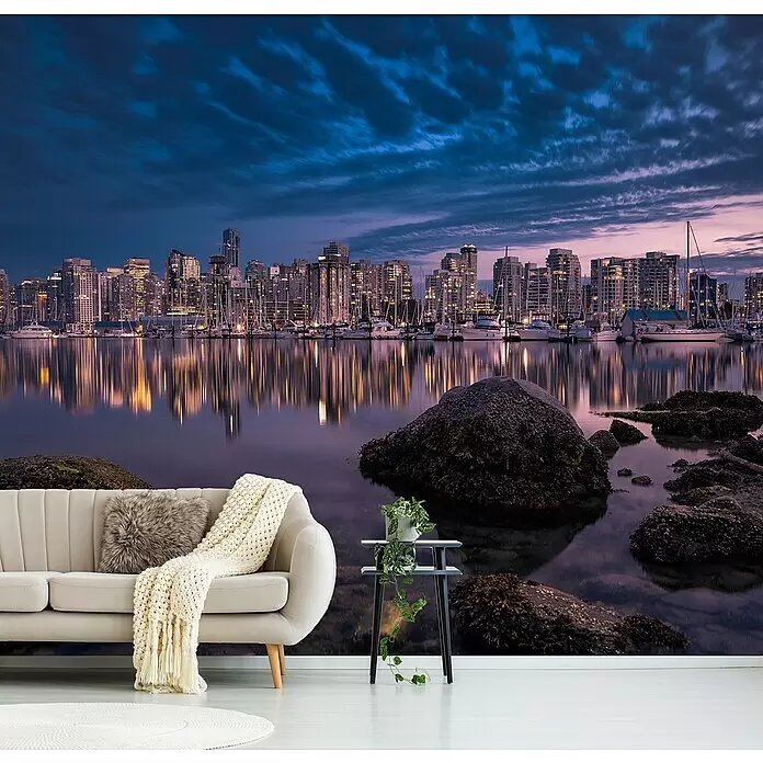 Papermoon Premium collection Fototapete Eine glühende Perle - B x H: 350 x 260 cm, Vlies - Wandbild mit Stadtsilhouette bei Dämmerung, im Wasser gespiegelt, mit Sofa und Pflanze im Vordergrund.