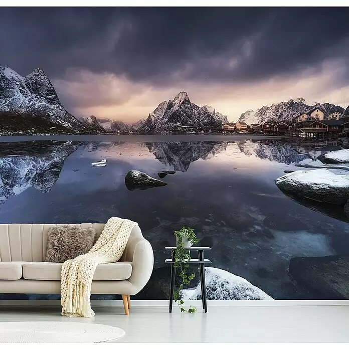 Papermoon Premium collection Fototapete Winterlich - B x H: 300 x 223 cm, Vlies - Wandbild einer verschneiten Berglandschaft mit spiegelndem See und Dorf unter dramatischem Himmel, in einem Wohnzimmer.
