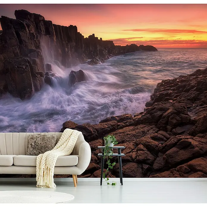Papermoon Premium collection Fototapete Wand am Meer - B x H: 450 x 280 cm, Vlies - Das Bild zeigt ein Wohnzimmer mit einer Felswandtapete, die eine dramatische Küstenlandschaft bei Sonnenuntergang darstellt. Eine stilvolle Couch und Beistelltisch ergänzen das Dekor.