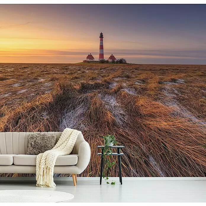 Papermoon Premium collection Fototapete Westerheversand - B x H: 250 x 186 cm, Vlies - Eine Wandtapete mit einem Leuchtturm in einem Feld bei Sonnenuntergang, als eindrucksvolle Kulisse hinter einem hellen Sofa und einem Beistelltisch im Wohnzimmer.