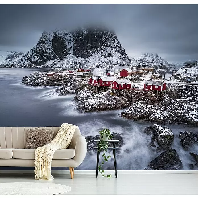Papermoon Premium collection Fototapete Lofoten Winter Nordlichter - B x H: 500 x 280 cm, Vlies - Eine Wandtapete zeigt eine verschneite norwegische Landschaft mit roten Fischerhütten am Meer, die eine ganze Wand hinter einem beigen Sofa bedeckt.