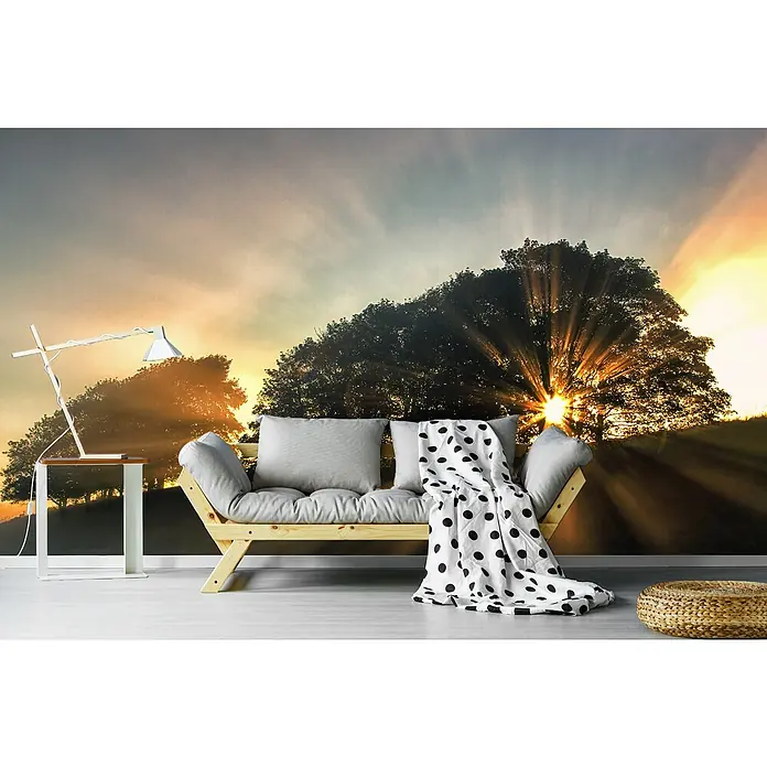 Papermoon Premium collection Fototapete Bäume Sonnenstr. - B x H: 350 x 260 cm, Vlies - Ein gemütliches Sofa mit Kissen und einer Decke mit Punktmuster steht vor einer Wandtapete mit Sonnenaufgang.
