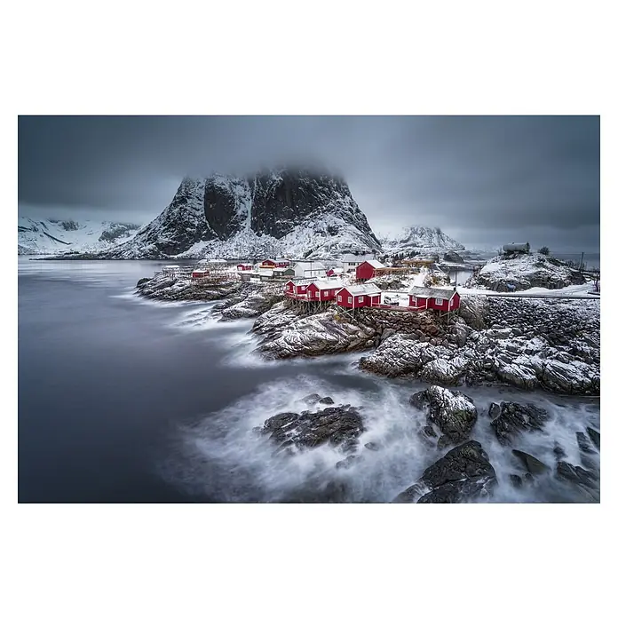 Papermoon Premium collection Fototapete Lofoten Winter Nordlichter - B x H: 500 x 280 cm, Vlies - Winterliche Landschaft mit traditionellen roten Fischerhütten auf schneebedeckten Felsen, umgeben von Meer und Bergen unter einem düsteren Himmel.