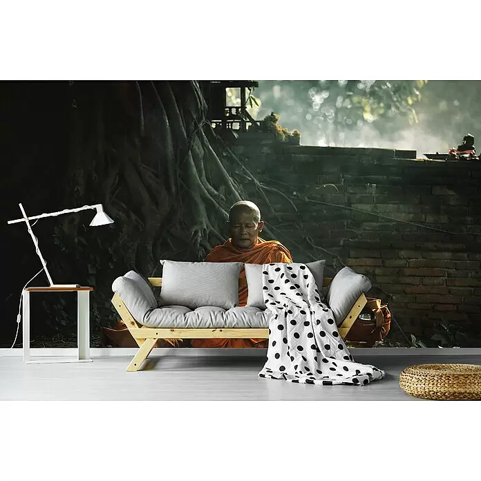 Papermoon Premium collection Fototapete Mönch morgendliche Meditation - B x H: 350 x 260 cm, Vlies - Holzsofa mit grauen Kissen und gepunkteter Decke, vor einem Wandbild eines meditierenden Mönchs.