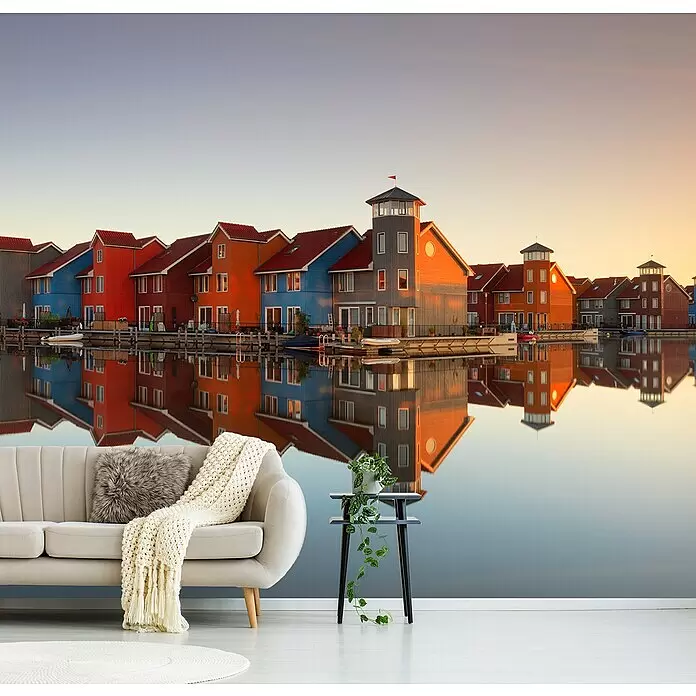Papermoon Premium collection Fototapete Buntes Dorf - B x H: 400 x 260 cm, Vlies - Gemütliches Wohnzimmer mit Wandbild farbiger Häuser, die sich bei Sonnenuntergang im Wasser spiegeln, dazu hellbeiges Sofa und Beistelltisch mit Pflanze.