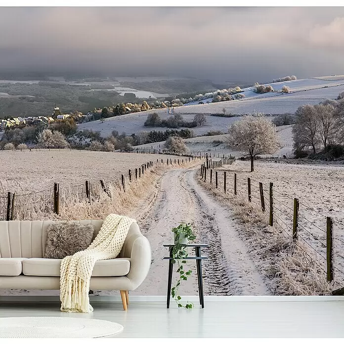 Papermoon Premium collection Fototapete Schneewanderung - B x H: 300 x 223 cm, Vlies - Ein gemütliches Zimmer mit beigem Sofa, Strickdecke, Kunstfellkissen, Beistelltisch mit Pflanze und cremefarbenem Teppich, vor einem Wandbild einer verschneiten Winterlandschaft.
