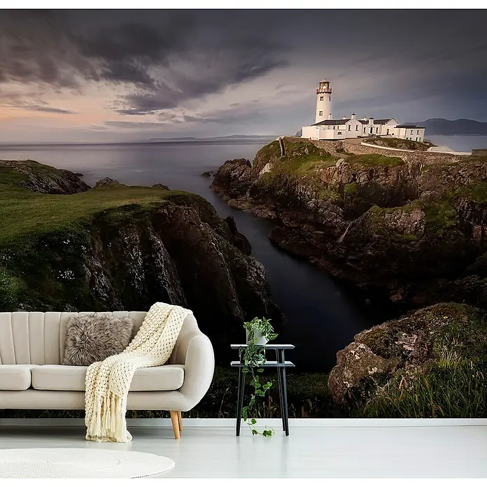 Papermoon Premium collection Fototapete Fanad Leuchtturm - B x H: 350 x 260 cm, Vlies - Wohnraum mit beigem Sofa, Hocker und einer Wandtapete, die einen Leuchtturm auf einer Klippe bei Sonnenuntergang zeigt.