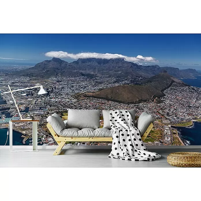 Papermoon Premium collection Fototapete Südafrika Kapstadt - B x H: 250 x 186 cm, Vlies - Ein modernes Futon-Sofa mit grauen Kissen, weißer Beistelltisch mit Lampe und geflochtener Pouf in Raum mit Stadtpanorama-Wandbild.