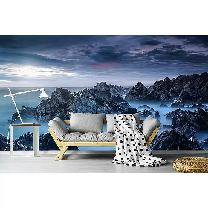 Papermoon Premium collection Fototapete Neblige Bergspitzen - B x H: 450 x 280 cm, Vlies - Holz-Futonsofa mit grauen Kissen und gepunkteter Decke in einem Raum mit Felsküsten-Wandbild.