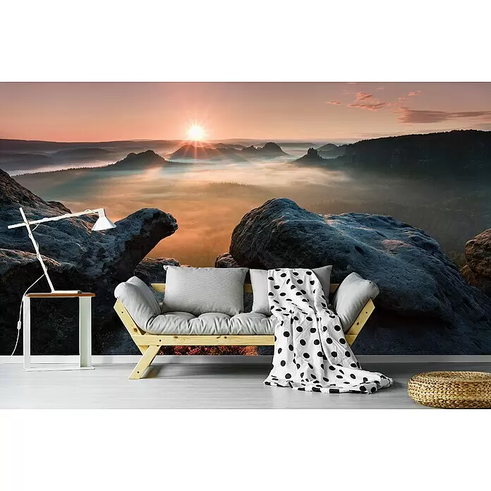 Papermoon Premium collection Fototapete Sonnenaufgang Felsen II - B x H: 300 x 223 cm, Vlies - Helles Holz-Futonsofa mit grauen Kissen und gepunkteter Decke in einem Raum mit Bergwandbild.