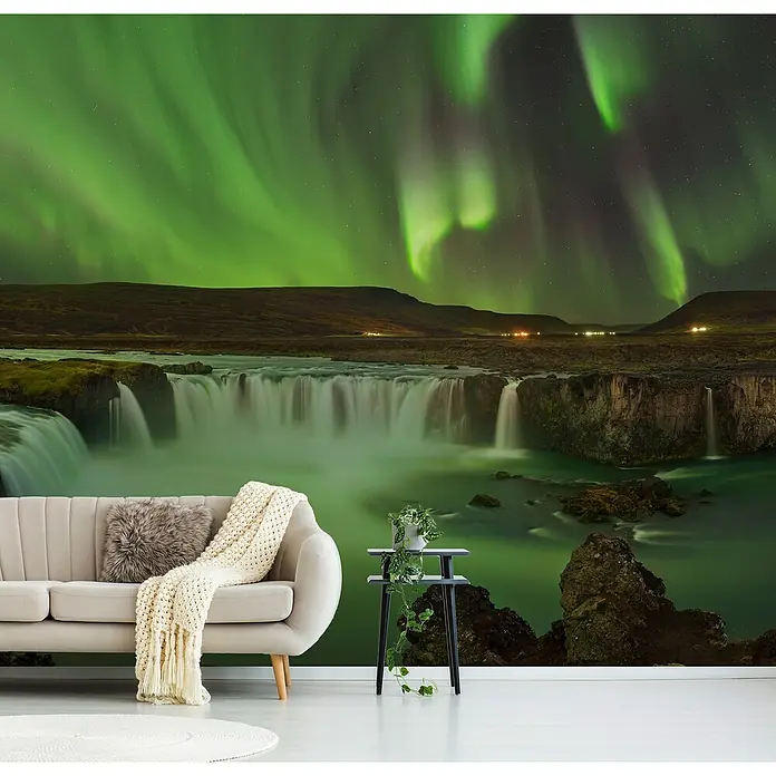 Papermoon Premium collection Fototapete Gödafoss Wasserfall Nordlichter - B x H: 300 x 223 cm, Vlies - Das Bild zeigt ein Sofa mit Kissen und Decke vor einem Wandbild mit Wasserfall und Nordlichtern, sowie einen Beistelltisch mit Pflanze.