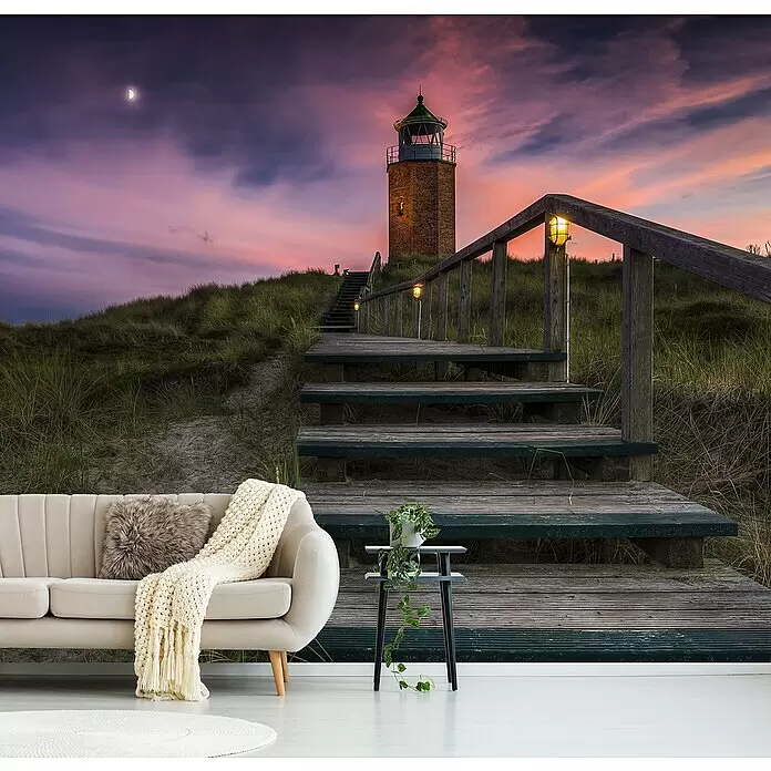 Papermoon Premium collection Fototapete Weg zum Leuchtturm - B x H: 250 x 186 cm, Vlies - Wandbild eines Leuchtturms und Dünen bei Sonnenuntergang. Ein beiges Sofa und Beistelltisch mit Pflanze sind im Vordergrund.
