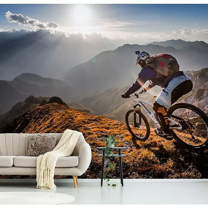 Papermoon Premium collection Fototapete Der Anruf des Berges - B x H: 400 x 260 cm, Vlies - Helles beiges Sofa mit Strickdecke und Fellkissen, und ein schwarzer Beistelltisch mit Pflanze, in einem Raum mit einem dramatischen Mountainbike-Wandbild.