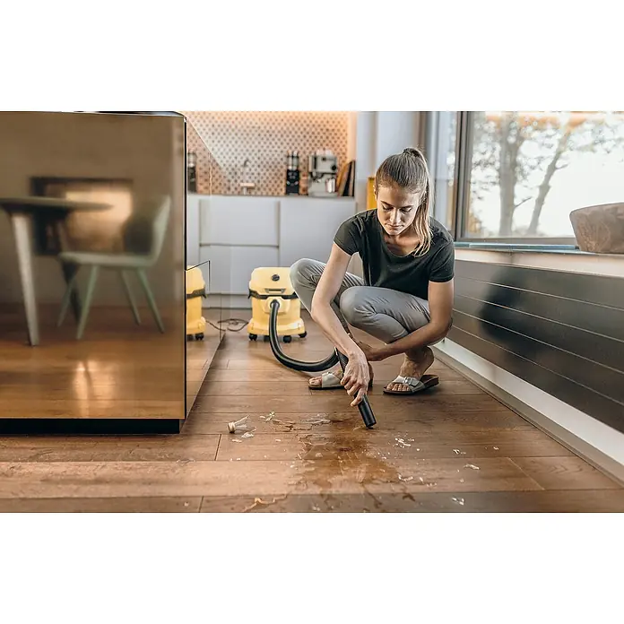 Kärcher Elektro-Nass- & Trockensauger WD 2 PLUS V-12/4/18 - 1.000 W, Behältervolumen: 12 l - Eine Frau reinigt mit einem gelben Nass-Trockensauger eine Flüssigkeit und Glasscherben auf einem Holzboden.