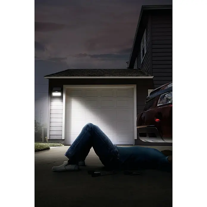 Ledvance LED-Solar-Außenwandleuchte Endura FloodSolarzelle, Kunststoff, Warmweiß, Dunkelgrau - Ein toter Mann liegt unter einem Auto vor einer Garage, möglicherweise beleuchtet durch eine Außenleuchte, was für Sicherheit sorgt.