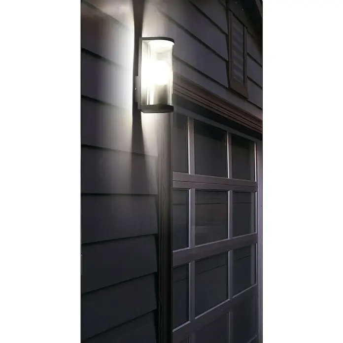 Ledvance Außenwandleuchte Endura Tbular - 11,7 x 8,2 x 22,2 cm, Dunkelgrau, IP44 - Nahaufnahme einer modernen Außenwandleuchte mit zylindrischem Schirm, die helles Licht an eine dunkle Fassade wirft.