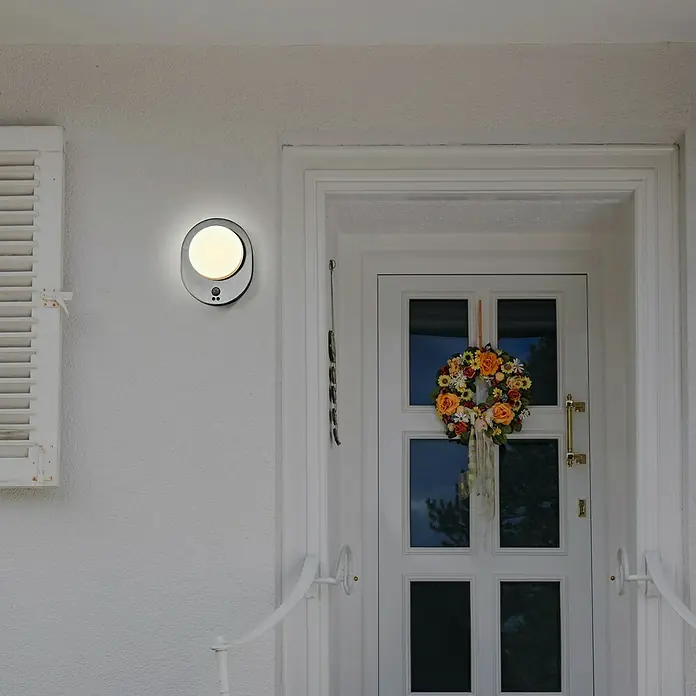 Osram LED-Außenwandleuchte Endura Disc Wall - 8 W, 24,4 x 20 x 6 cm, Silber, IP44 - Eine moderne, runde LED-Wandleuchte in Schwarz und Weiß, die neben einer Haustür mit einem Blumenkranz hängt und für stilvolle Beleuchtung sorgt.