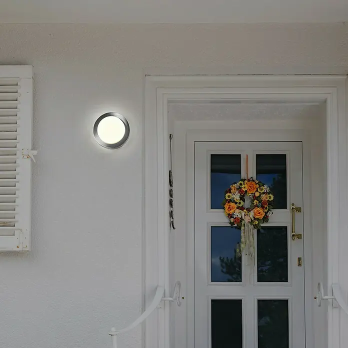Osram LED-Außenwandleuchte Endura Disc Wall - 8 W, 20 x 20 x 6 cm, Silber, IP44 - Nahaufnahme einer modernen runden Wandleuchte mit Metalleinfassung, die an einer weißen Außenwand neben einer Haustür befestigt ist.