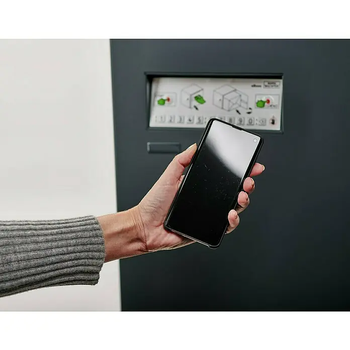 Burg-Wächter Paketbox eBoxx GV 645 - L x B x H: 45 x 57,5 x 76 cm, Anthrazit, Stahl, Namensschild, Fassungsvermögen: 197 l - Eine Person hält ein schwarzes Smartphone vor einem grauen Automaten mit einer Bedienungsanleitung. Der Fokus liegt auf dem modernen Mobiltelefon.