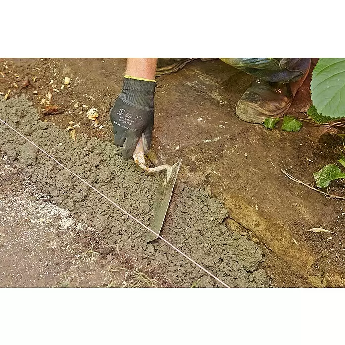 Quick-Mix Gartenbaubeton mit Trass - 30 kg - Schwarzer Arbeitshandschuh an einer Hand, die mit einer Kelle Beton entlang einer rosa Richtschnur im Graben verteilt.