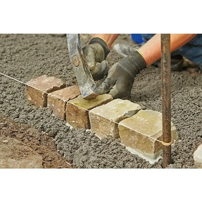Quick-Mix Drainagemörtel - 25 kg - Nahaufnahme eines Pflasterers, der Pflastersteine mit einem Hammer entlang einer Richtschnur verlegt. Die Pflastersteine werden sorgfältig ausgerichtet.