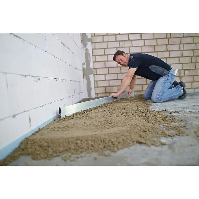 Quick-Mix Estrichbeton B03 - 40 kg - Ein Mann kniet und glättet mit einer Metallschiene eine Sandschicht auf einem Boden, um ihn für weitere Bauarbeiten vorzubereiten.