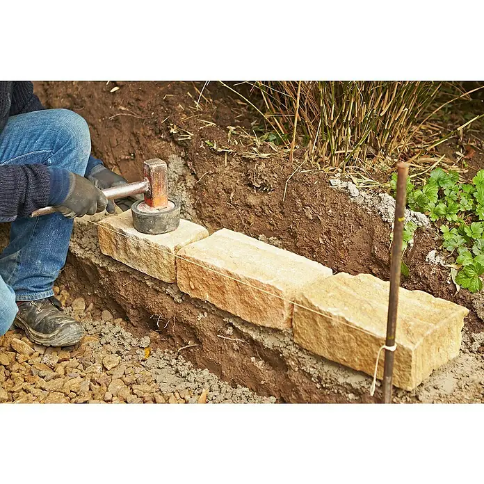 Quick-Mix Gartenmauermörtel - 30 kg - Ein Arbeiter verlegt sorgfältig Sandsteine für eine Gartenmauer, wobei er einen Gummihammer benutzt, um die Steine auszurichten.