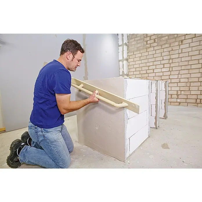 Quick-Mix Hagalith Kalkzement-Haftputz FF - 40 kg - Ein Mann kniet, während er mit einer langen, flachen Latte Putz auf eine Wand aus Porenbetonsteinen aufträgt, um die Oberfläche zu ebnen.