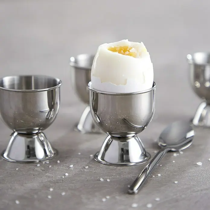 Set de huevera para mesa - Ø x Al: 4,5 x 5 cm, 4 ud., Metal - Una escena de desayuno con un huevo pasado por agua en una huevera de acero inoxidable, acompañado de una cuchara y una pizca de sal. Las hueveras son de metal brillante.
