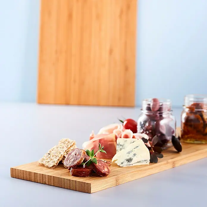 Tabla de cortar con mango - 16 x 61 x 1,5 cm, Bambú - Salchichas, quesos, galletas saladas y aceitunas en conserva están dispuestos en una tabla de bambú, con otra tabla de bambú en blanco al fondo.