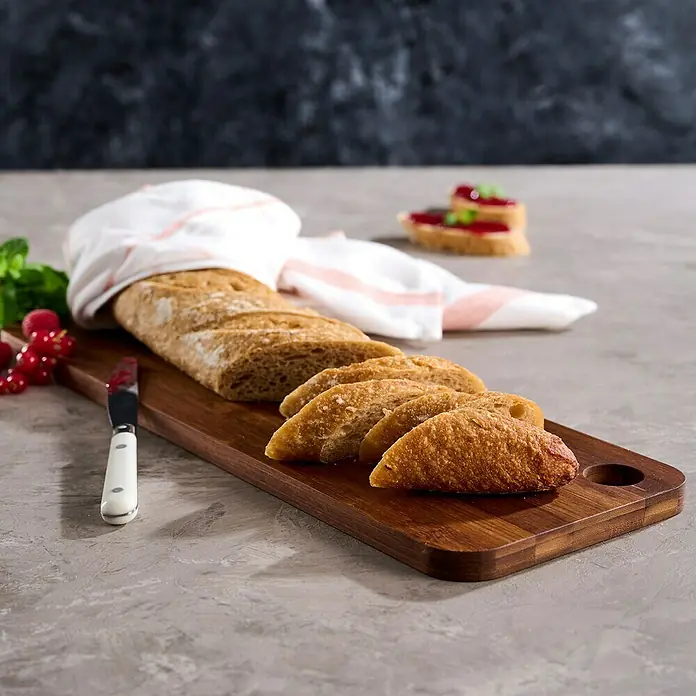 Tabla de cortar - 15 x 45 x 1,5 cm, Madera - Una baguette rebanada yace sobre una tabla de madera cubierta con un paño, junto con un cuchillo y grosellas.