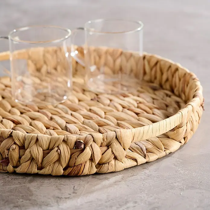 Bandeja natural - Diámetro: 32 cm, Fibra natural, Natural - Primer plano de una bandeja redonda de servir tejida con dos vasos transparentes, destacando la elegancia rústica de la bandeja.