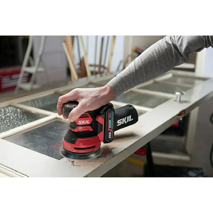 Skil PWRCore 20V Akku-Exzenterschleifer 3745 CA - 20 V, Ohne Akku, Durchmesser Schleifteller: 125 mm - Ein Mann schleift eine Tür mit einem SKIL Exzenterschleifer. Das Gerät ist rot-schwarz und kabellos.