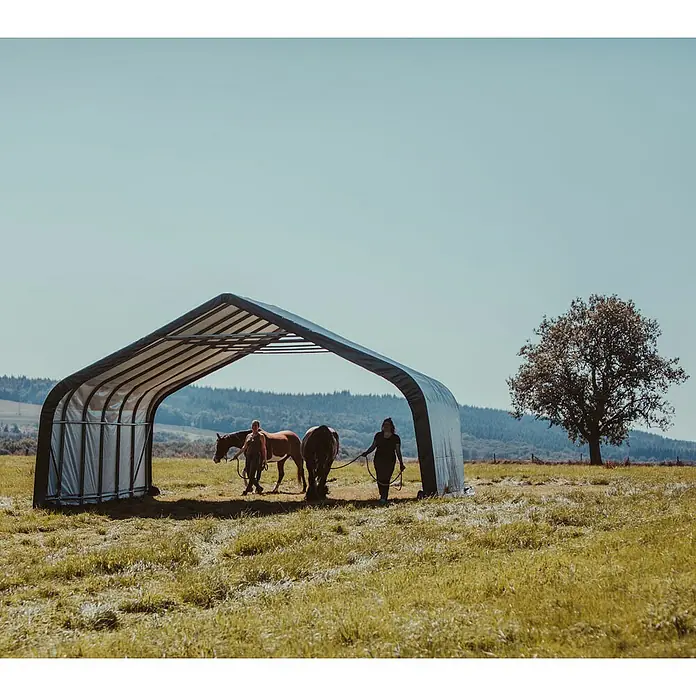 ShelterLogic Garage - 730 x 680 x 380 cm, Stahl - Pferdestall aus Stahlgewebe mit zwei Pferden und zwei Personen auf einer Wiese unter strahlend blauem Himmel.