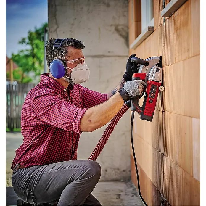 Einhell Rozadora TE-MA 1500 - 1.500 W, Diámetro disco: 125 mm - Hombre usando una rozadora de pared Einhell en una pared de ladrillo, con gafas de seguridad, máscara y protección auditiva.