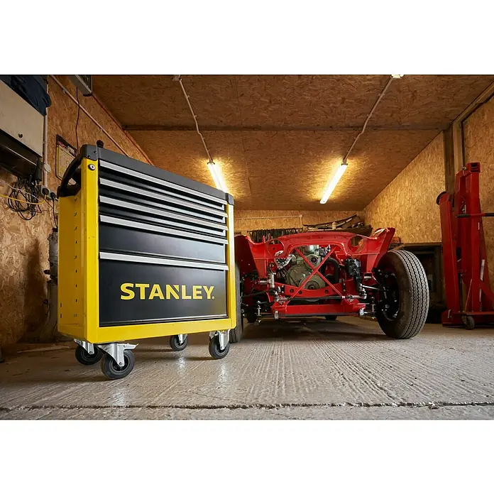 Stanley Carro portaherramientas STST74305-1 - L x An x Al: 67,6 x 42,6 x 85,9 cm, Número de cajones: 5 ud., Capacidad de carga: 300 kg - Imagen de taller con un carro de herramientas Stanley amarillo y negro frente a un chasis de coche rojo en un garaje.