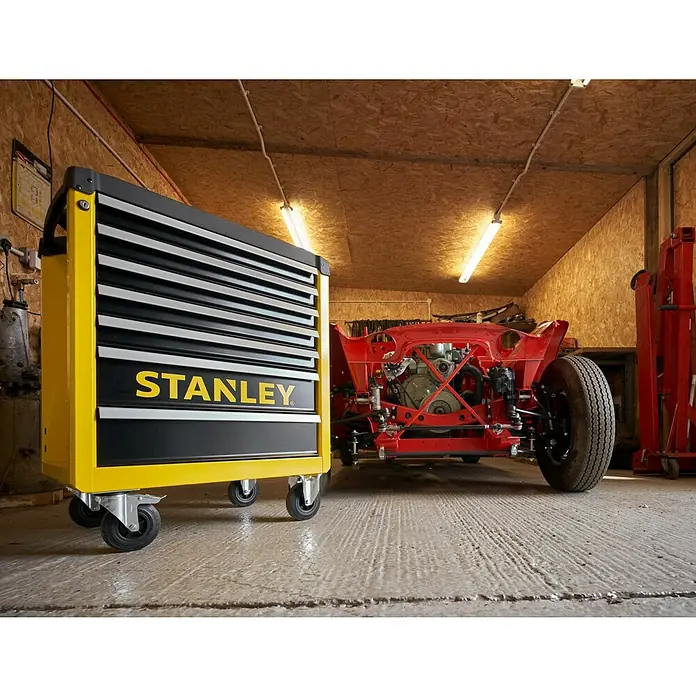 Stanley Carro portaherramientas STST74306-1 - L x An x Al: 74 x 45,9 x 89,8 cm, Número de cajones: 7 ud., Capacidad de carga: 300 kg - Carro de herramientas Stanley en un taller con un coche rojo al fondo. Almacenamiento de herramientas práctico para cada artesano.