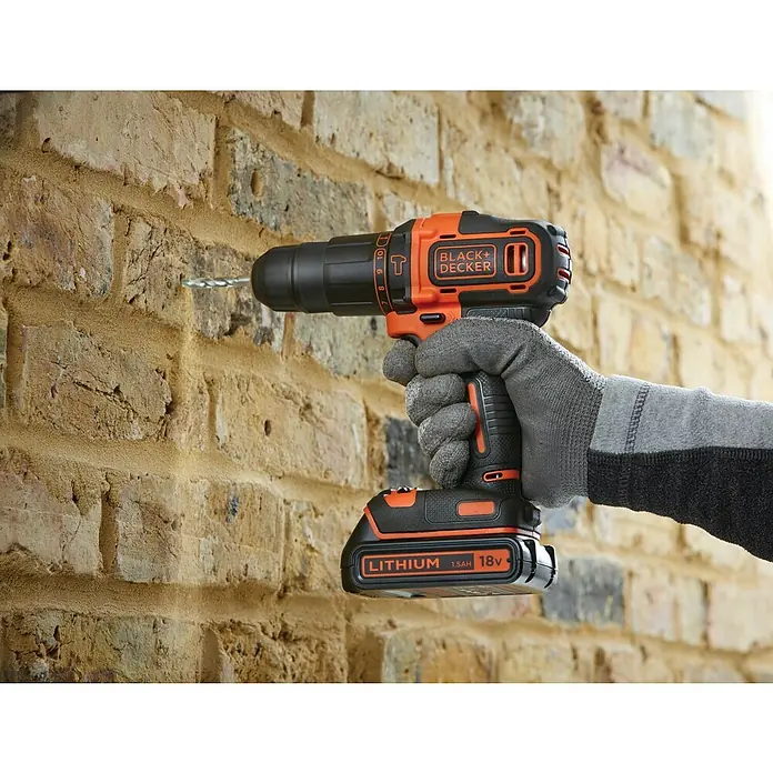 Black+Decker Taladro atornillador percutor de batería BDCHD18KB2 - 18 V, 2 baterías, 2 Ah, 40 Nm - Un taladro inalámbrico Black+Decker perfora una pared de ladrillo, sujeto por una mano con guante.